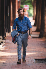 Man walking down city sidewalk with jacket in hand. He is wearing a blue dress shirt and grey pants.