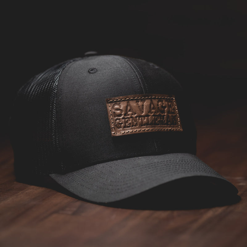 Savage Gentleman Leather Patch Trucker Hat in black mesh and black.